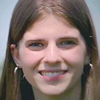 Headshot of guest Rylee Nelson. They are smiling warmly at the camera, with long straight brown hair and circular hoop earrings, photographed outdoors with a soft blurred background.