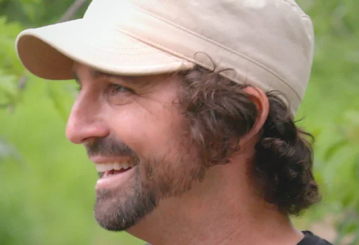 Headshot of guest Brian Counselman. They are smiling and looking to the side, with dark curly hair pulled back and a beard, wearing a tan baseball cap, photographed outdoors with a soft green background.
