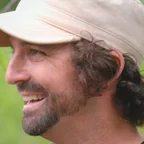 Headshot of guest Brian Counselman. They are smiling and looking to the side, with dark curly hair pulled back and a beard, wearing a tan baseball cap, photographed outdoors with a soft green background.