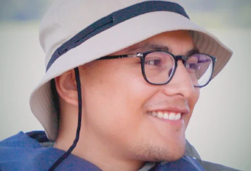 Headshot of guest David Ortiz. They are smiling and looking to the side, wearing a tan bucket hat with a chin strap and black-framed glasses, photographed outdoors with a soft blurred background.