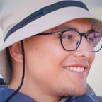 Headshot of guest David Ortiz. They are smiling and looking to the side, wearing a tan bucket hat with a chin strap and black-framed glasses, photographed outdoors with a soft blurred background.