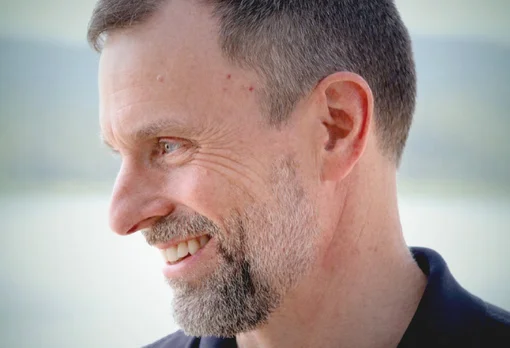 Headshot of guest Paul Dearlove. They are smiling and looking to the side, with short grey hair and a short beard, wearing a dark shirt, photographed with a soft light background.
