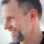 Headshot of guest Paul Dearlove. They are smiling and looking to the side, with short grey hair and a short beard, wearing a dark shirt, photographed with a soft light background.