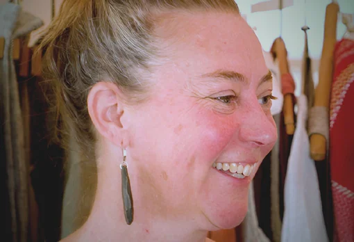 Headshot of guest Leslie Schroeder. They are smiling and looking to the side, with blonde hair pulled up, wearing long drop earrings, photographed indoors with clothing racks visible in the background.