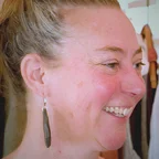 Headshot of guest Leslie Schroeder. They are smiling and looking to the side, with blonde hair pulled up, wearing long drop earrings, photographed indoors with clothing racks visible in the background.