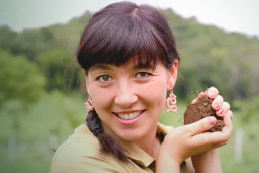 A person with dark hair in a braid smiles at the camera while holding a clump of rich brown soil. They wear worm-shaped earrings and a khaki shirt, standing outdoors with a green, hilly landscape in the background.