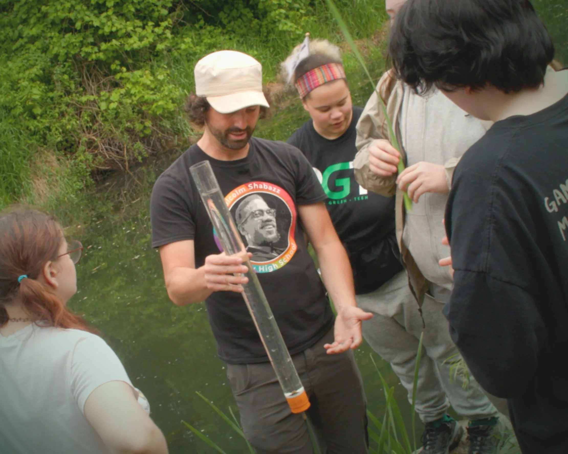 An adult in a white cap and black graphic t-shirt holds a long clear cylindrical water sampling tube while explaining it to a small group of young people gathered along the edge of a pond. One young person wears a colorful headband and a green 