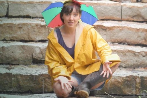 A person wearing a yellow rain slicker and a small multicolored umbrella hat sits on wet stone steps in the rain, looking directly at the camera with a playful expression. They wear rubber boots and a purple top beneath the open raincoat.