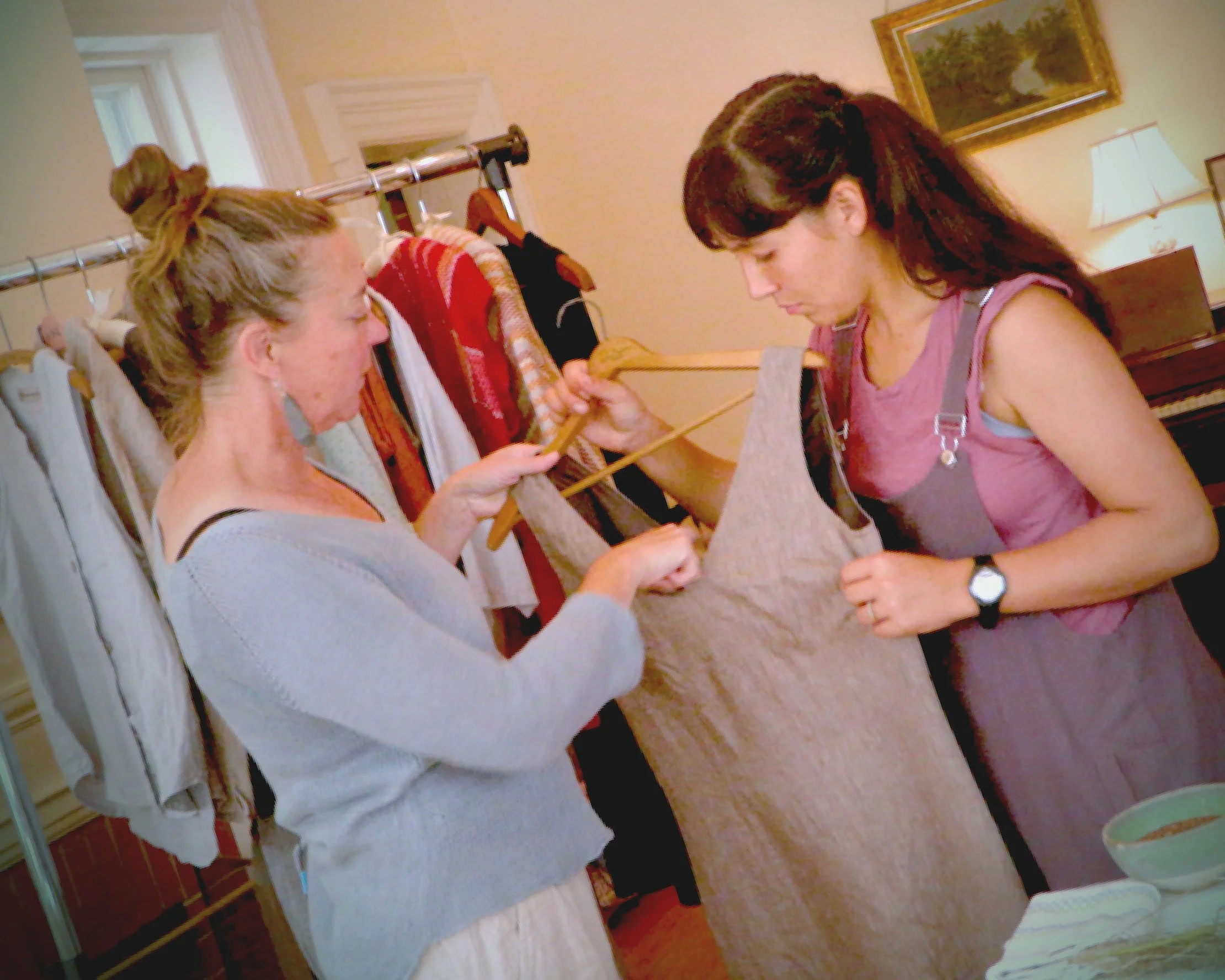 Two people stand indoors examining a linen garment on a wooden hanger. One person, wearing a light gray sweater with their hair up, holds out the hanger while the other, in a mauve sleeveless top, inspects the fabric closely. A clothing rack with various garments hangs in the background.