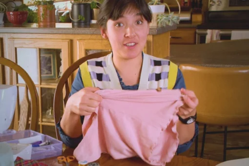 A person with pulled back dark hair and bangs sits at a wooden table, holding up a crumpled pink fabric garment with a wide-eyed expression. They wear a colorful patterned vest over a blue shirt. Sewing supplies and scissors are visible on the table in front of them.
