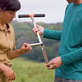 Two people stand in a green field examining a T-shaped soil probe. One person wears a tan jumpsuit; the other wears a teal long-sleeve shirt. Rolling hills are visible in the background.