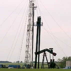 An oil drilling rig and pump jack stand in a flat, open field under an overcast sky. Several vehicles are parked nearby.