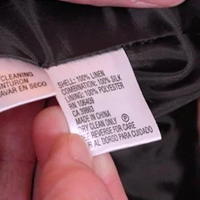 A hand holds up a white fabric care label on a dark garment, showing fabric content and care instructions including 