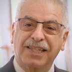 Headshot of guest Majid Sarmadi. They are smiling warmly at the camera, with short white hair, a grey mustache, and thin-framed glasses, wearing a suit jacket, photographed indoors with a soft light background.