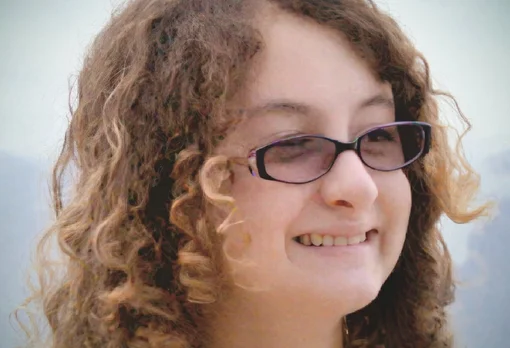 Headshot of guest Stella Gomez Phillips. They are smiling and looking slightly to the side, with curly brown hair and purple-framed glasses, photographed outdoors with a soft light background.