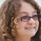Headshot of guest Stella Gomez Phillips. They are smiling and looking slightly to the side, with curly brown hair and purple-framed glasses, photographed outdoors with a soft light background.
