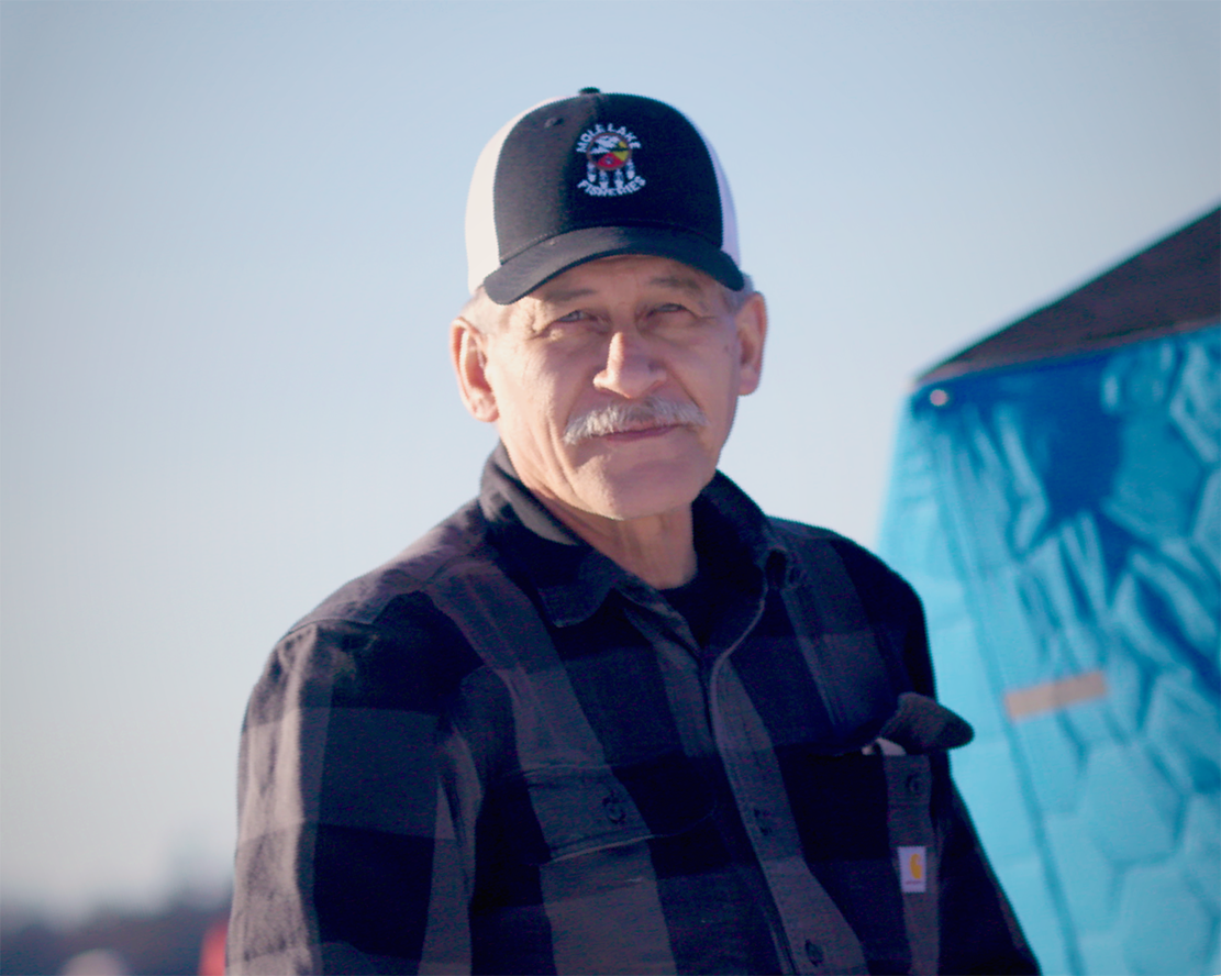 A man wearing a Mole Lake Fisheries cap and plaid flannel shirt stands outdoors, with a blue ice fishing shelter visible behind him.