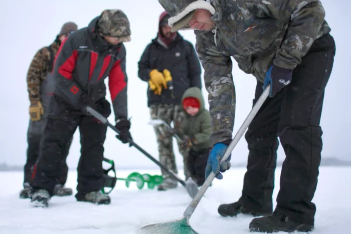 A group of adults and a child work together on a frozen lake, clearing snow and drilling holes with an ice auger during a winter fishing outing.