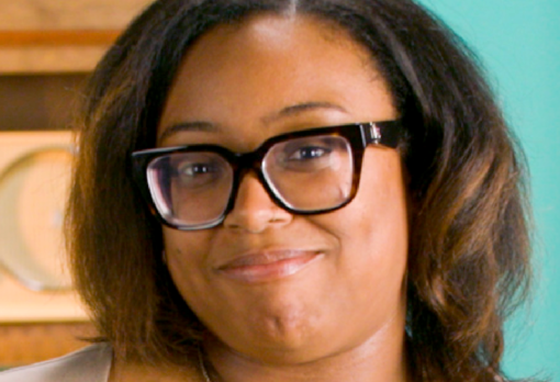 Person with dark curly hair and black-framed glasses smiles slightly, in front of a teal studio wall.