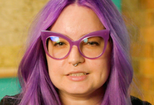 Person with bright purple hair and purple-framed glasses looks at the camera, brick wall behind them.