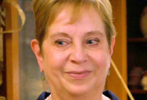 Person with short blond hair smiles slightly, with shelving of artifacts visible behind them.