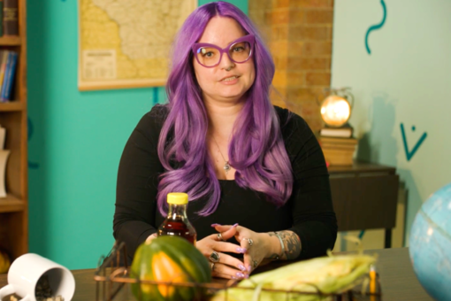 Person with purple hair at a desk with squash, corn, a bottle of dark liquid, and a globe behind them.