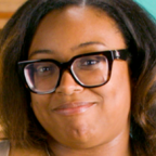 Person with dark curly hair and black-framed glasses smiles slightly, in front of a teal studio wall.