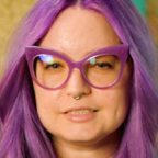 Person with bright purple hair and purple-framed glasses looks at the camera, brick wall behind them.