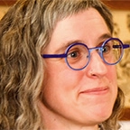 Close-up portrait of a woman with curly gray-streaked hair and round blue-framed glasses, smiling slightly, with tattoo flash art visible on the wall behind her.