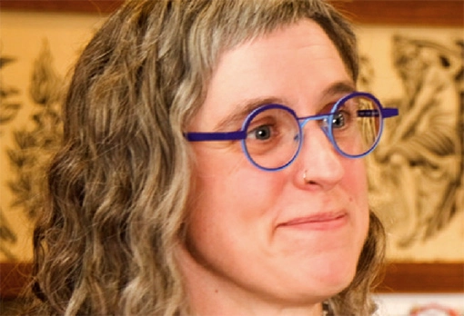 Close-up portrait of a woman with curly gray-streaked hair and round blue-framed glasses, smiling slightly, with tattoo flash art visible on the wall behind her.
