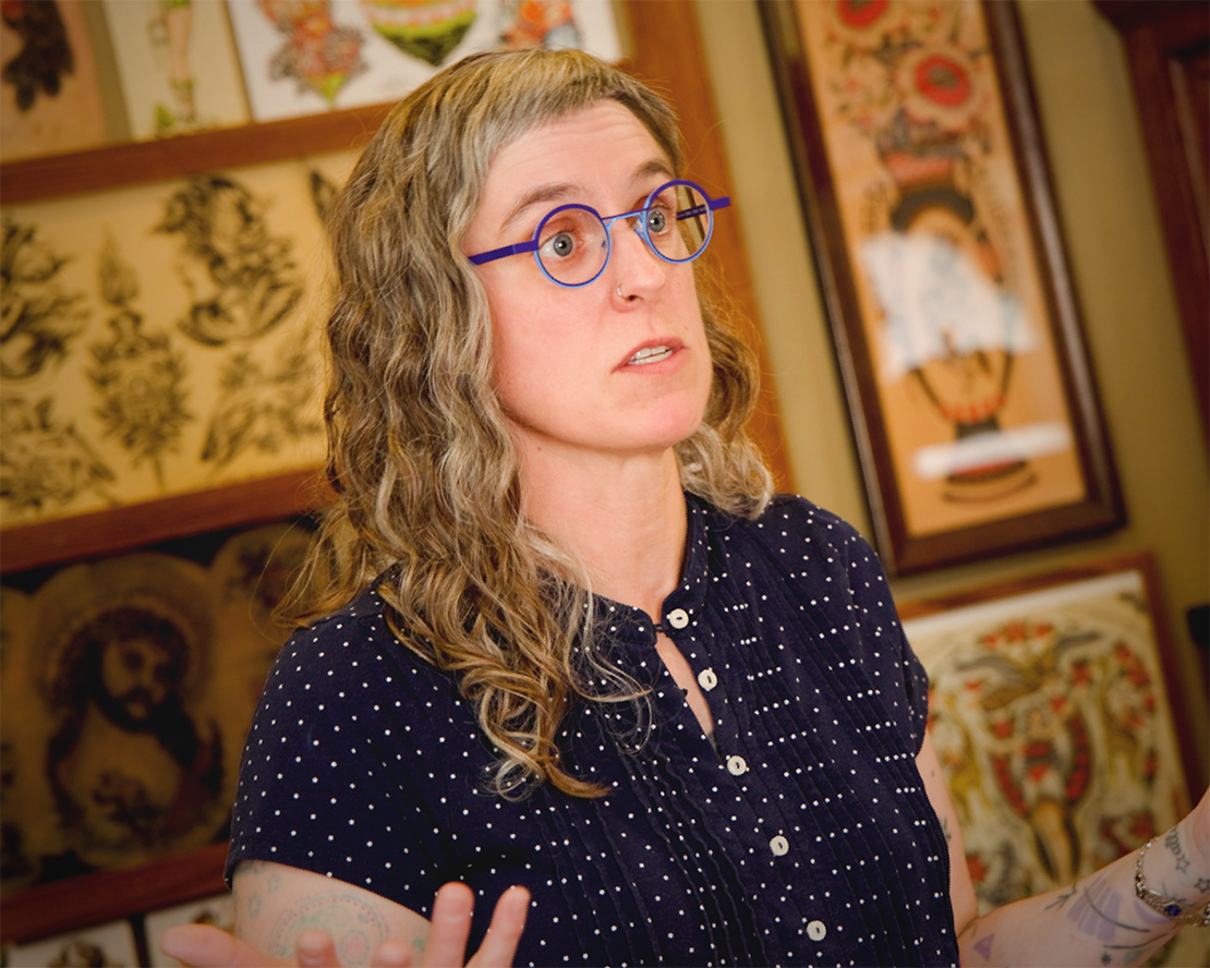 Woman with curly gray-blonde hair and round blue glasses speaks while gesturing with her hands, wearing a navy polka-dot blouse, framed by vintage tattoo flash art on the wall behind her.