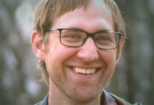 Headshot of Chris Wilson, a smiling man with short brown hair, glasses, and a beard, wearing a gray shirt.