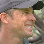 Profile view of Evan Larson, a smiling man with a low ponytail wearing a gray baseball cap and teal shirt, outdoors with trees in the background.
