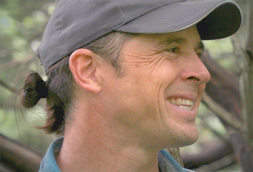 Profile view of Evan Larson, a smiling man with a low ponytail wearing a gray baseball cap and teal shirt, outdoors with trees in the background.