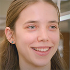 Close-up of Evelyn Zadzilka, a smiling young woman with long brown hair and a small teal earring, indoors.