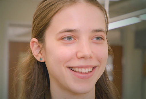 Close-up of Evelyn Zadzilka, a smiling young woman with long brown hair and a small teal earring, indoors.