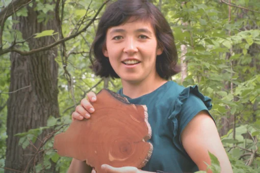 A smiling woman holds a cross-cut log slice showing its growth rings, standing outdoors in a wooded area.
