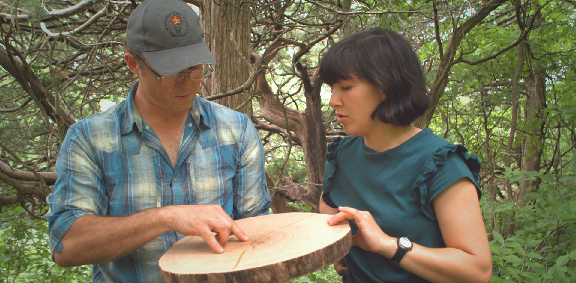 A man in a cap and plaid shirt points to growth rings on a cut log while a woman looks on, both standing outdoors surrounded by trees.