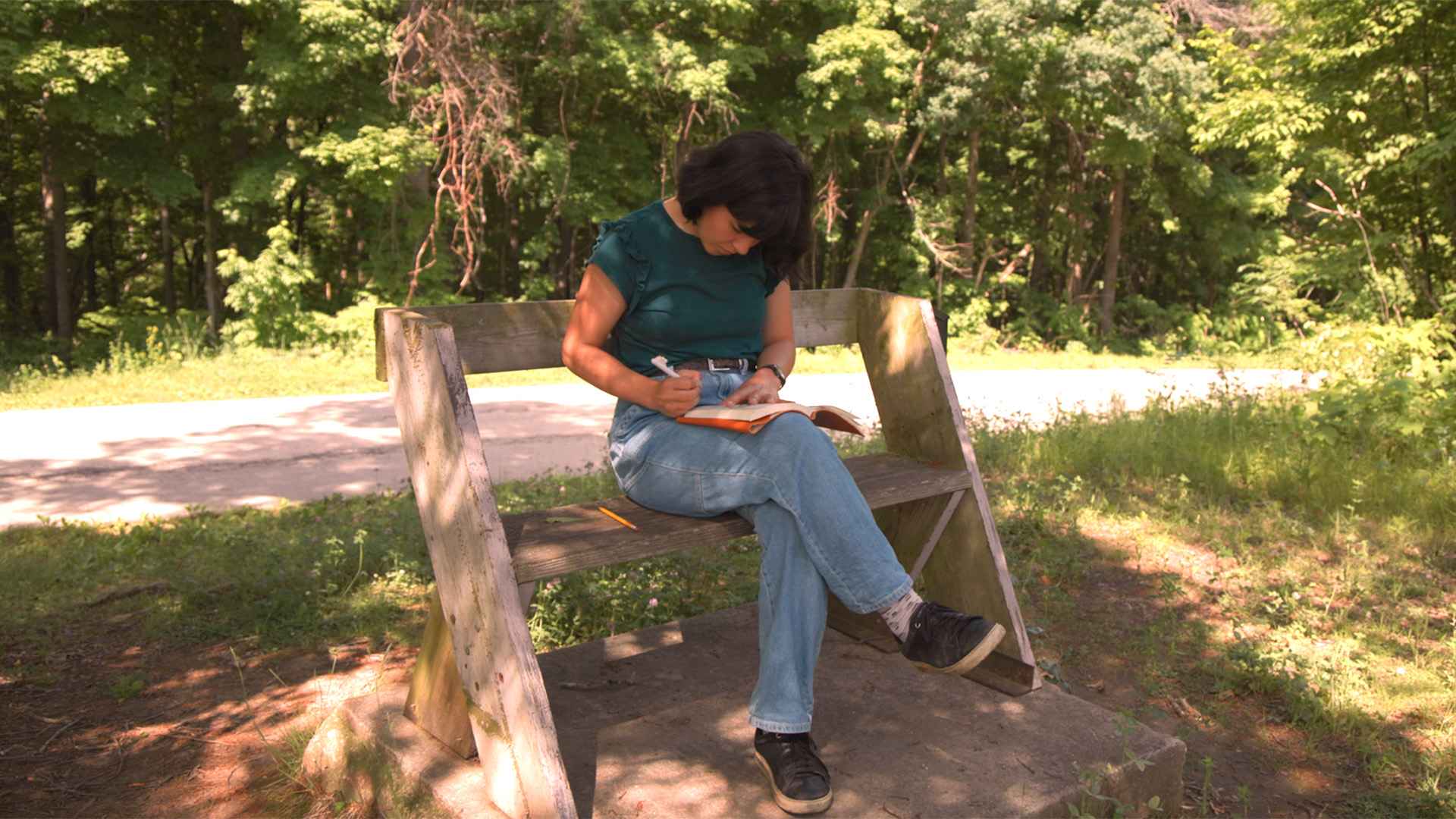 A woman sits on a rustic wooden bench beside a trail, writing in a notebook, surrounded by dense green forest.