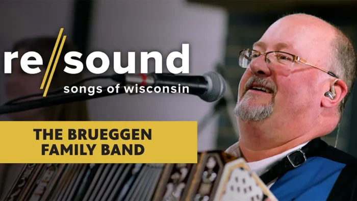 Promotional graphic reading "re/sound: songs of wisconsin" and "The Brueggen Family Band." A smiling Gary Brueggen with glasses and a goatee sings into a microphone while playing an ornate concertina. He wears an in-ear monitor, and a blurred bandmate is behind him.