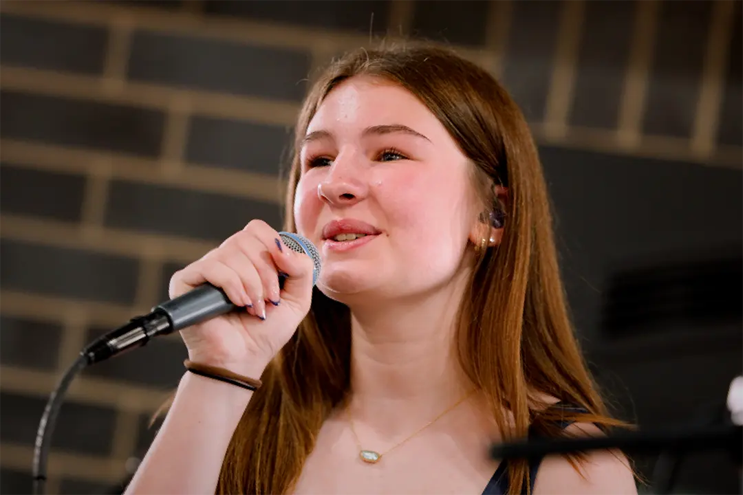 Matti Brueggen with long brown hair smiling as she sings into a microphone during a performance