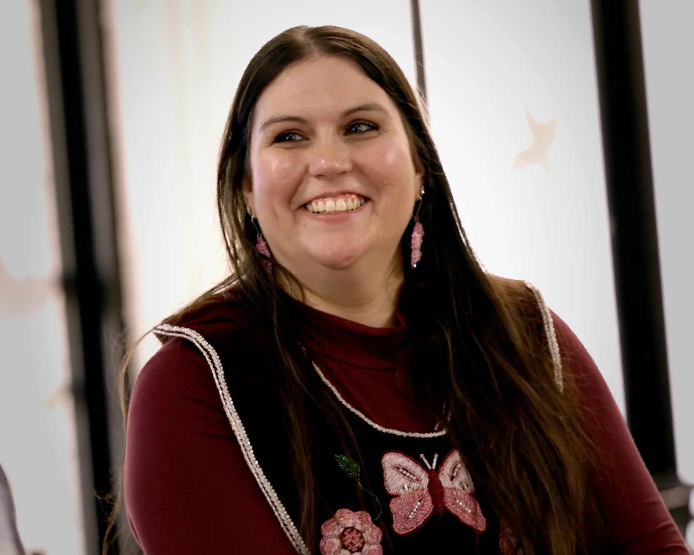 Smiling woman with long dark hair, beaded earrings, and a black vest beaded with a pink butterfly and flower.