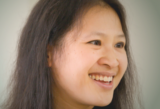 Smiling person with long dark hair looking off to the side, in front of a pale wall.