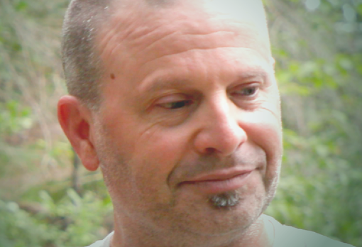 Person with close-cropped hair and a small goatee smiling slightly, with green foliage behind.