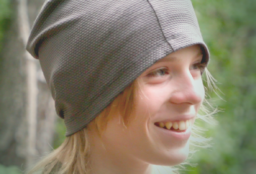Smiling person in a gray knit cap with blond hair, looking off to the side in a wooded setting.