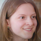 Smiling person with long light brown hair and a small green earring, indoors.