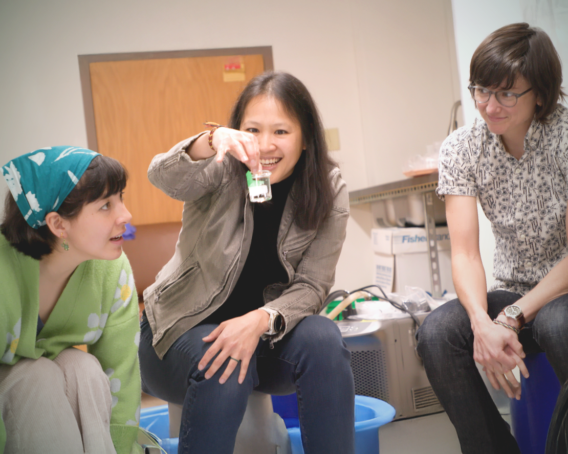 In a lab, a smiling person holds up a small beaker with a specimen as two people seated nearby watch.